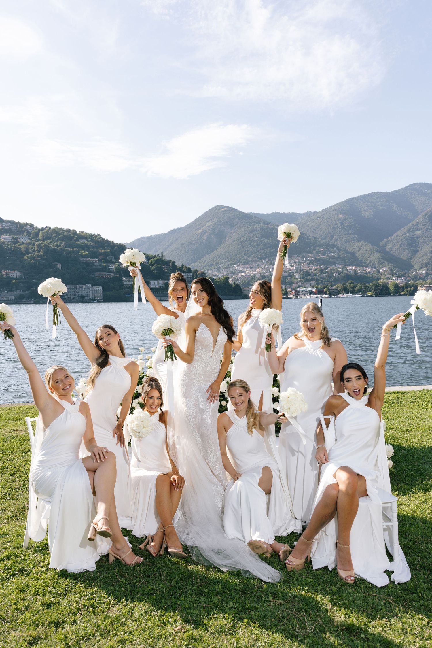 A group in flowing white dresses celebrate joyfully with bouquets by a scenic lakeside mountain landscape in Italy.
