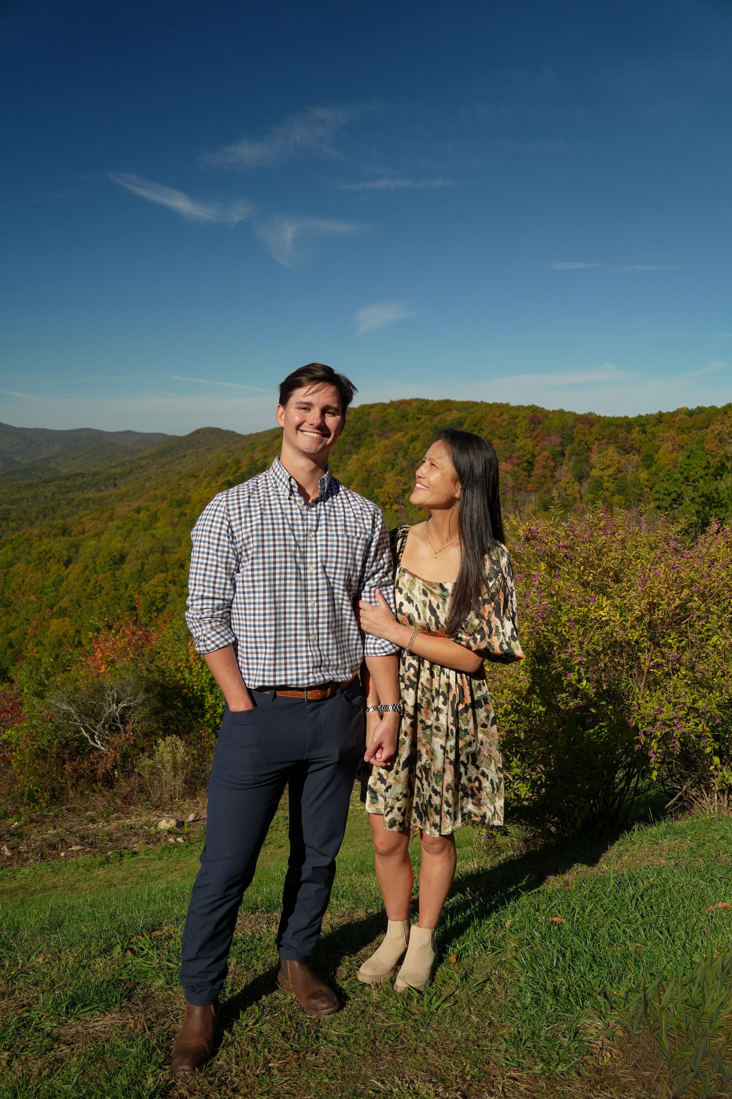 Couple posing on a scenic mountain overlook with colorful autumn foliage under a bright blue sky.