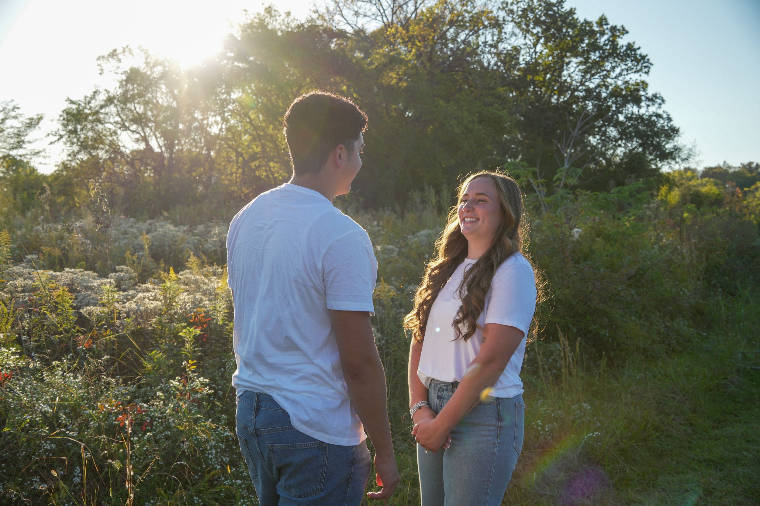 Couple smiling at each other in a sunlit outdoor field surrounded by lush green trees and wildflowers.
