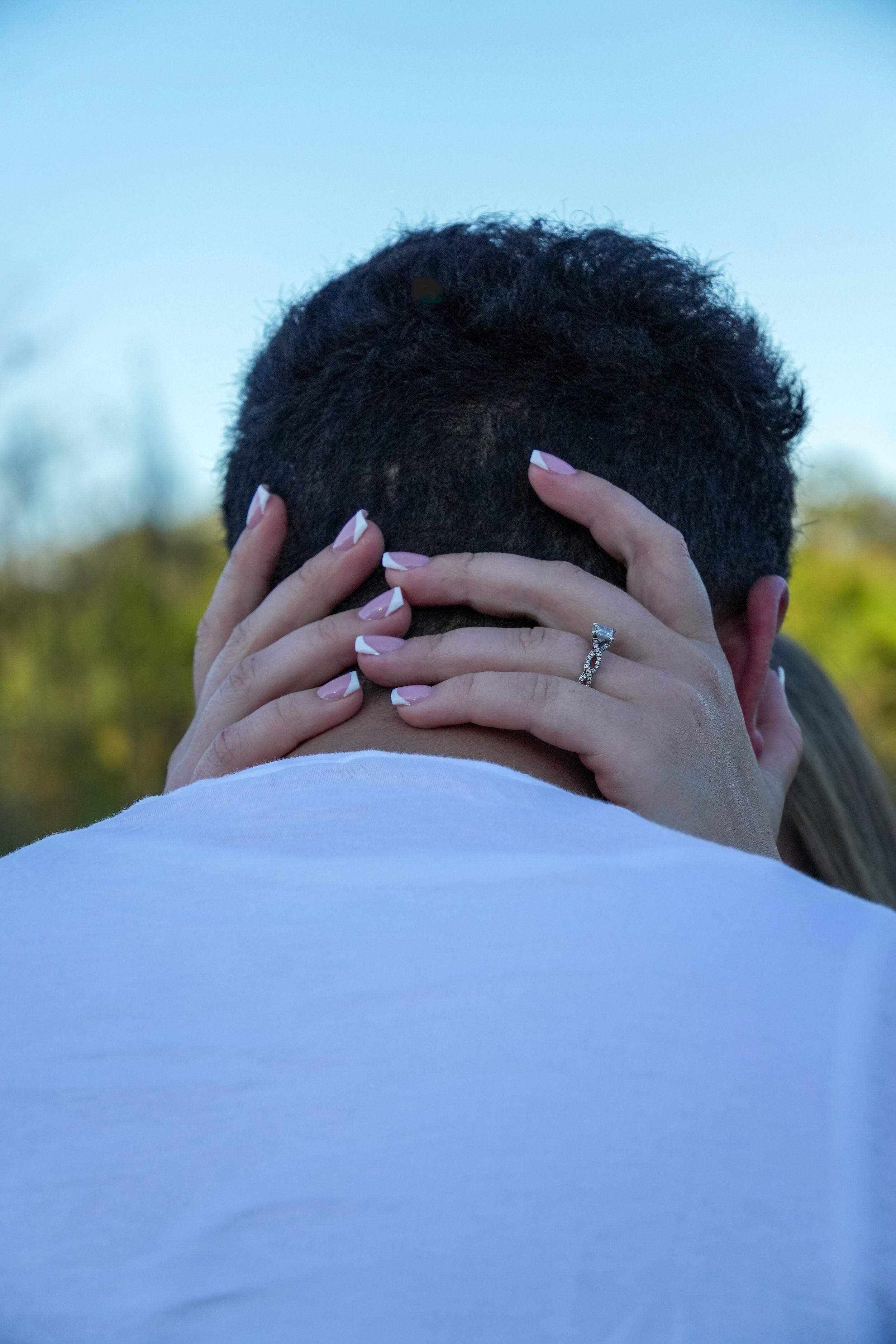 Couple embracing outdoors, woman's hands with engagement ring resting on man's neck, blue sky background.