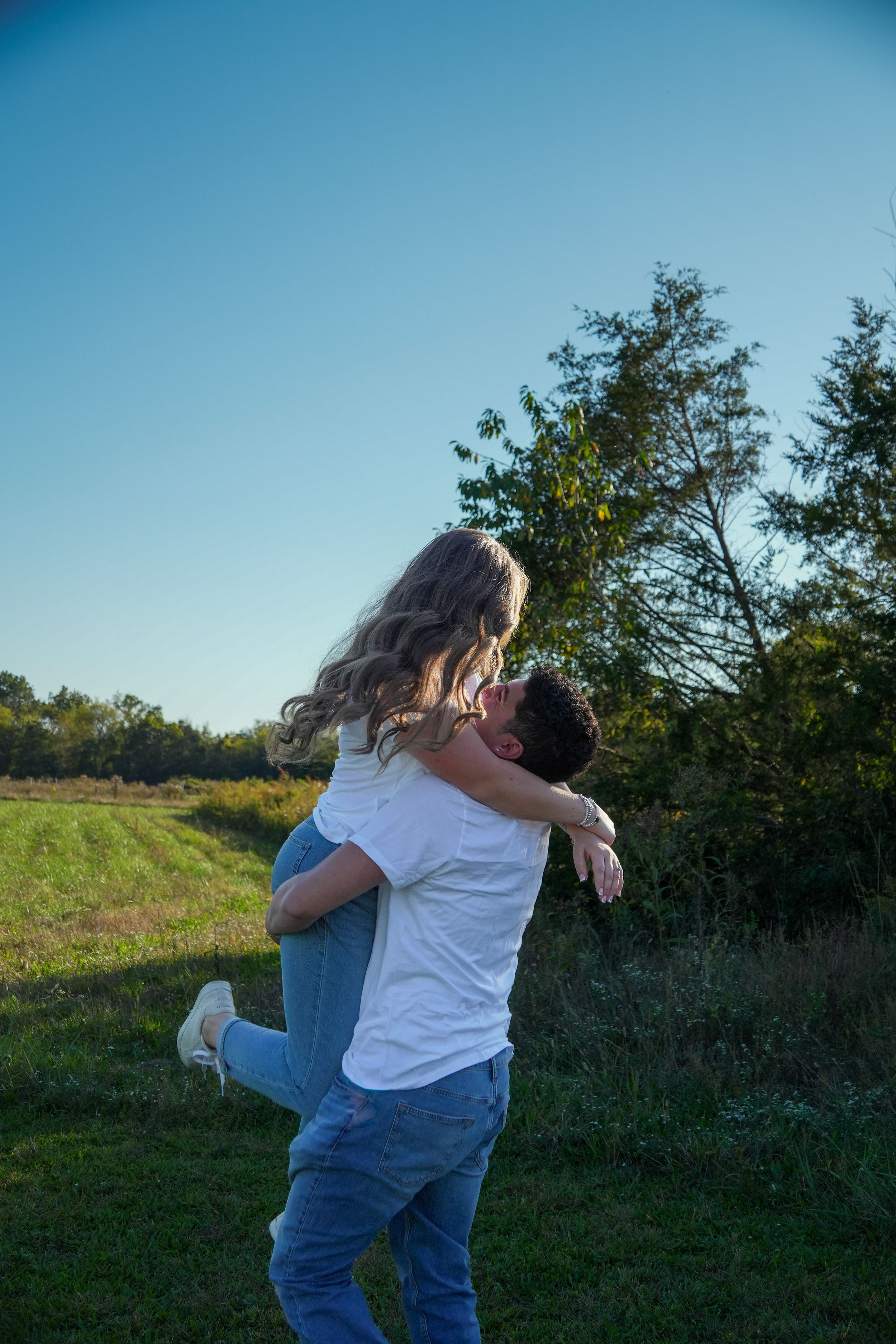 A couple embracing outdoors, man lifting woman in a sunny green field with trees and blue sky backdrop.