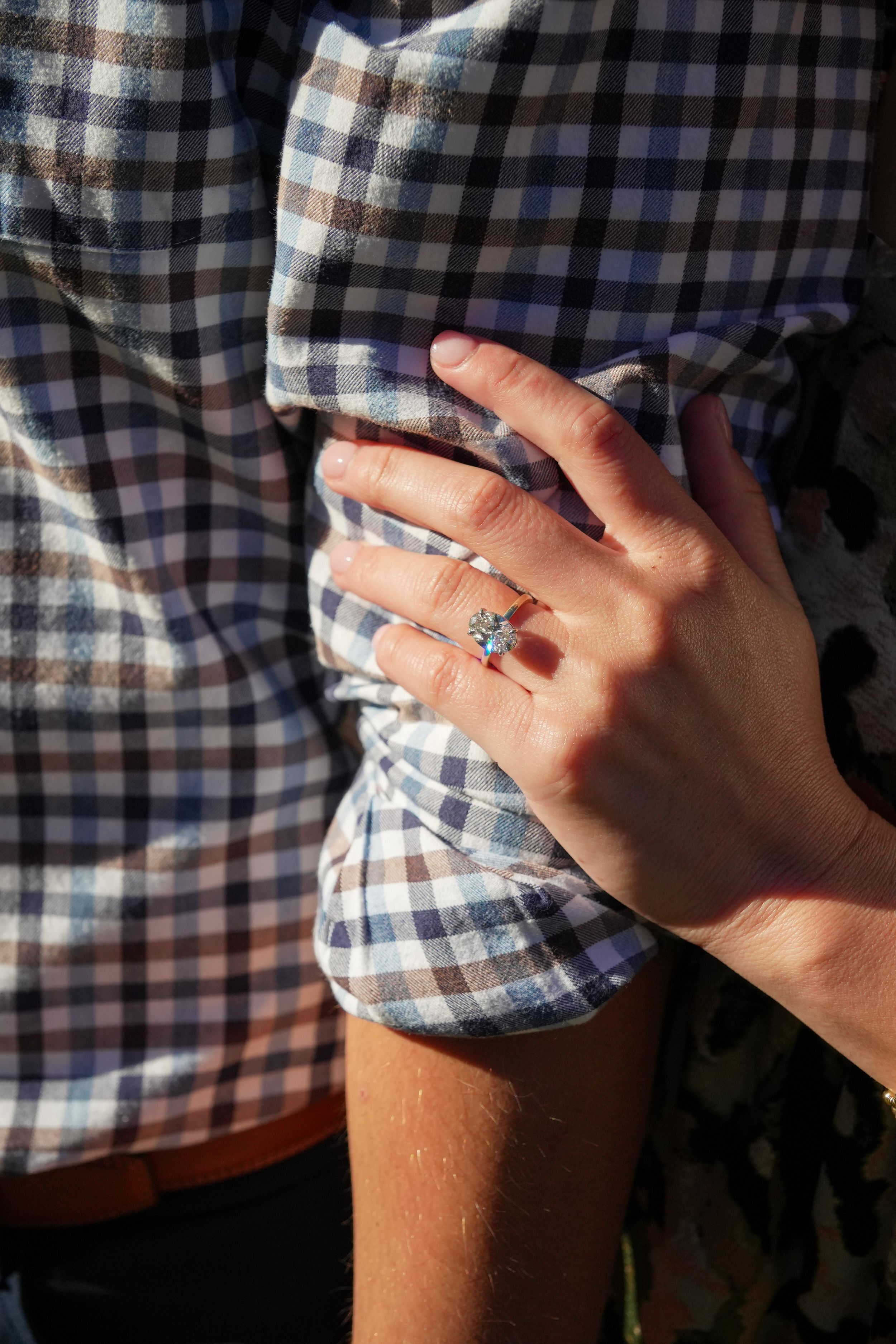 Close-up of a hand wearing a diamond engagement ring resting on a man's blue plaid flannel shirt.