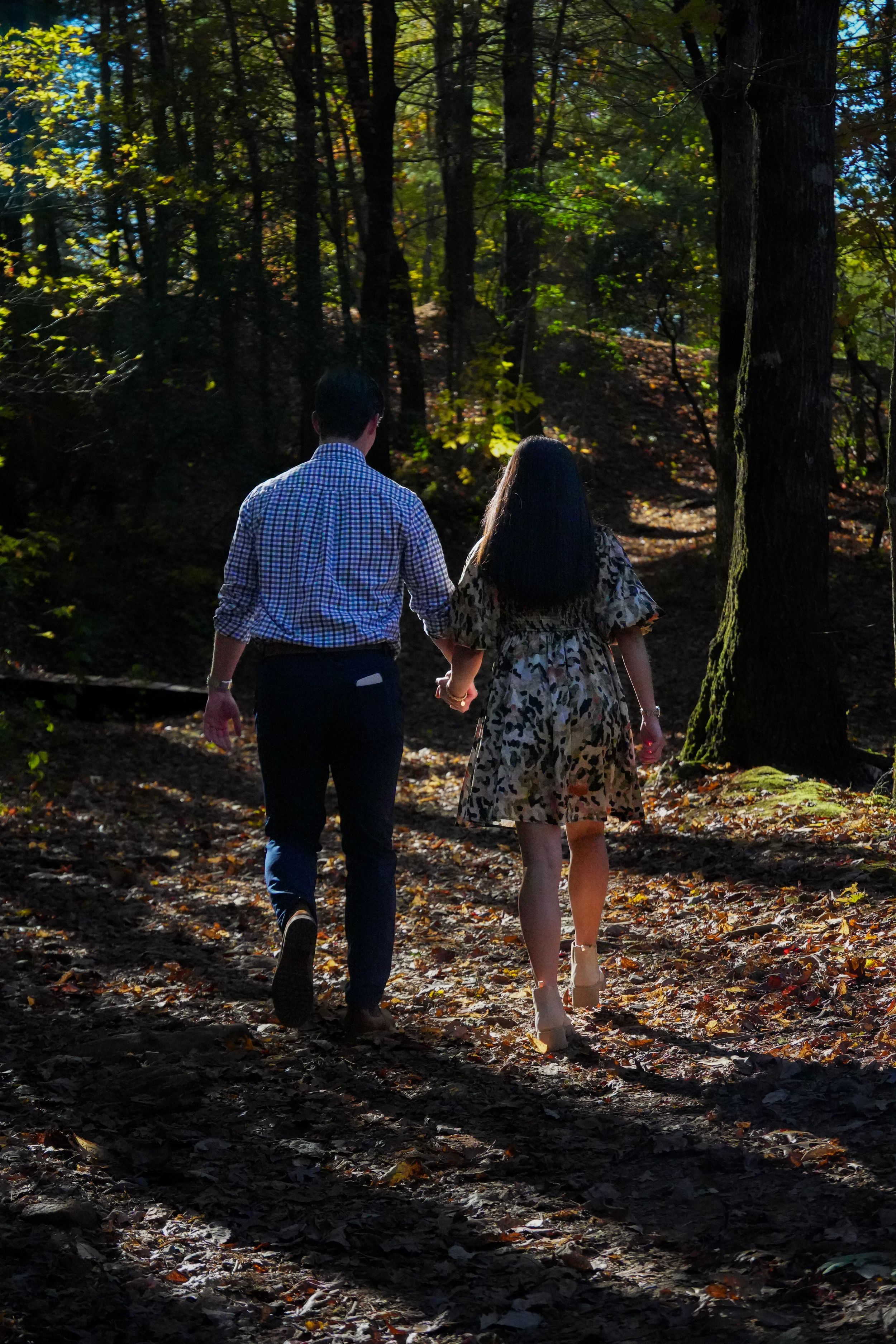 Couple holding hands walking through a sunlit autumn forest with fallen leaves on the ground.