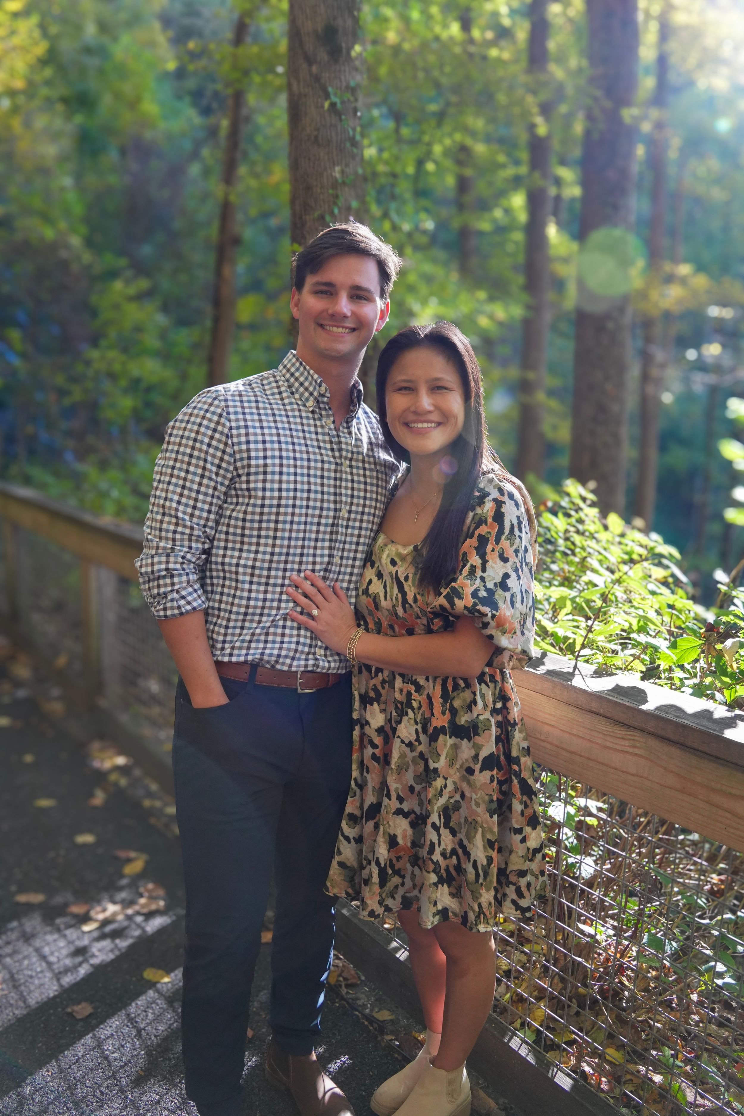 Smiling couple posing outdoors on a wooden bridge surrounded by lush green trees in natural sunlight.