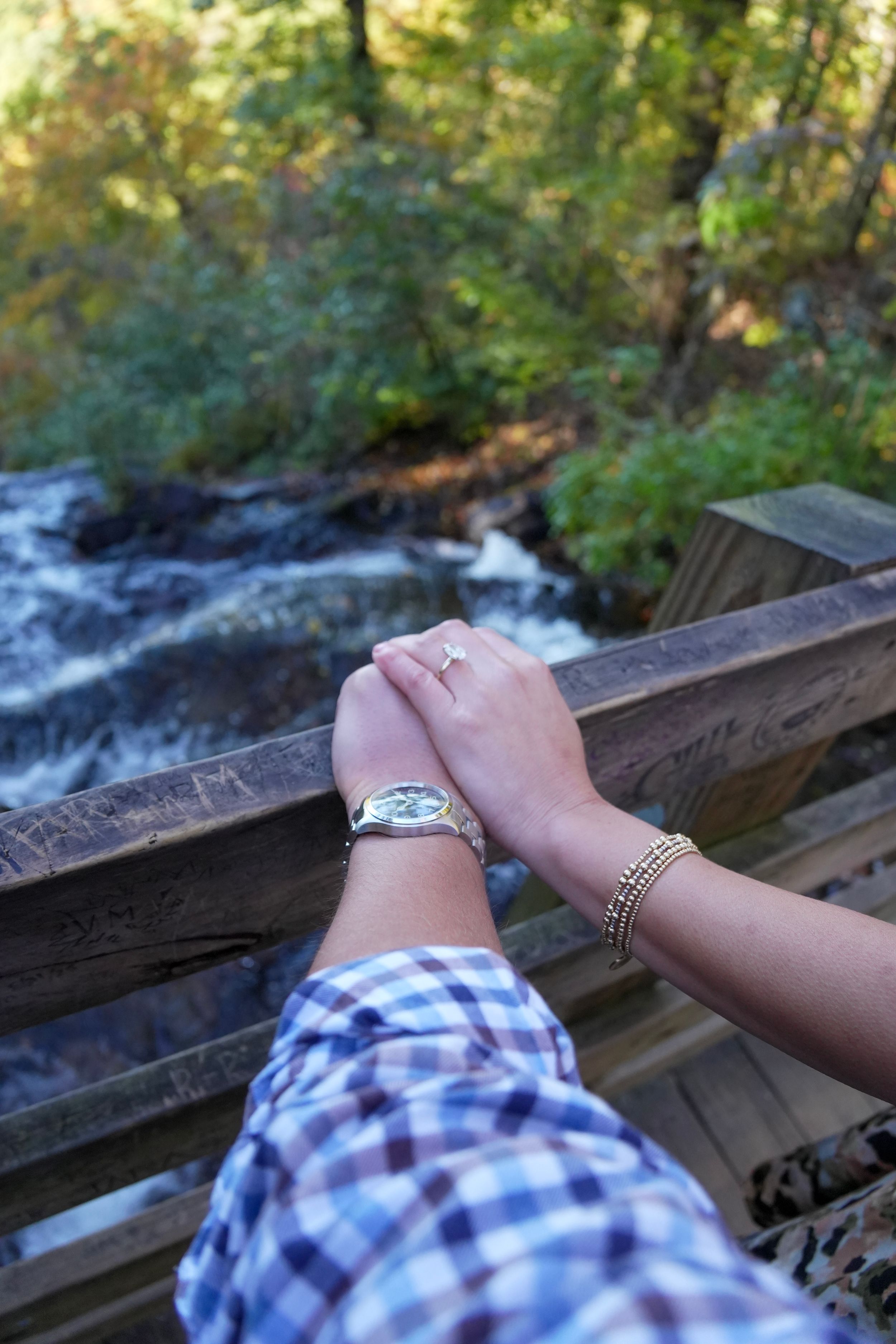 Couple holding hands on a wooden bridge overlooking a forest stream, one wearing a watch and ring.