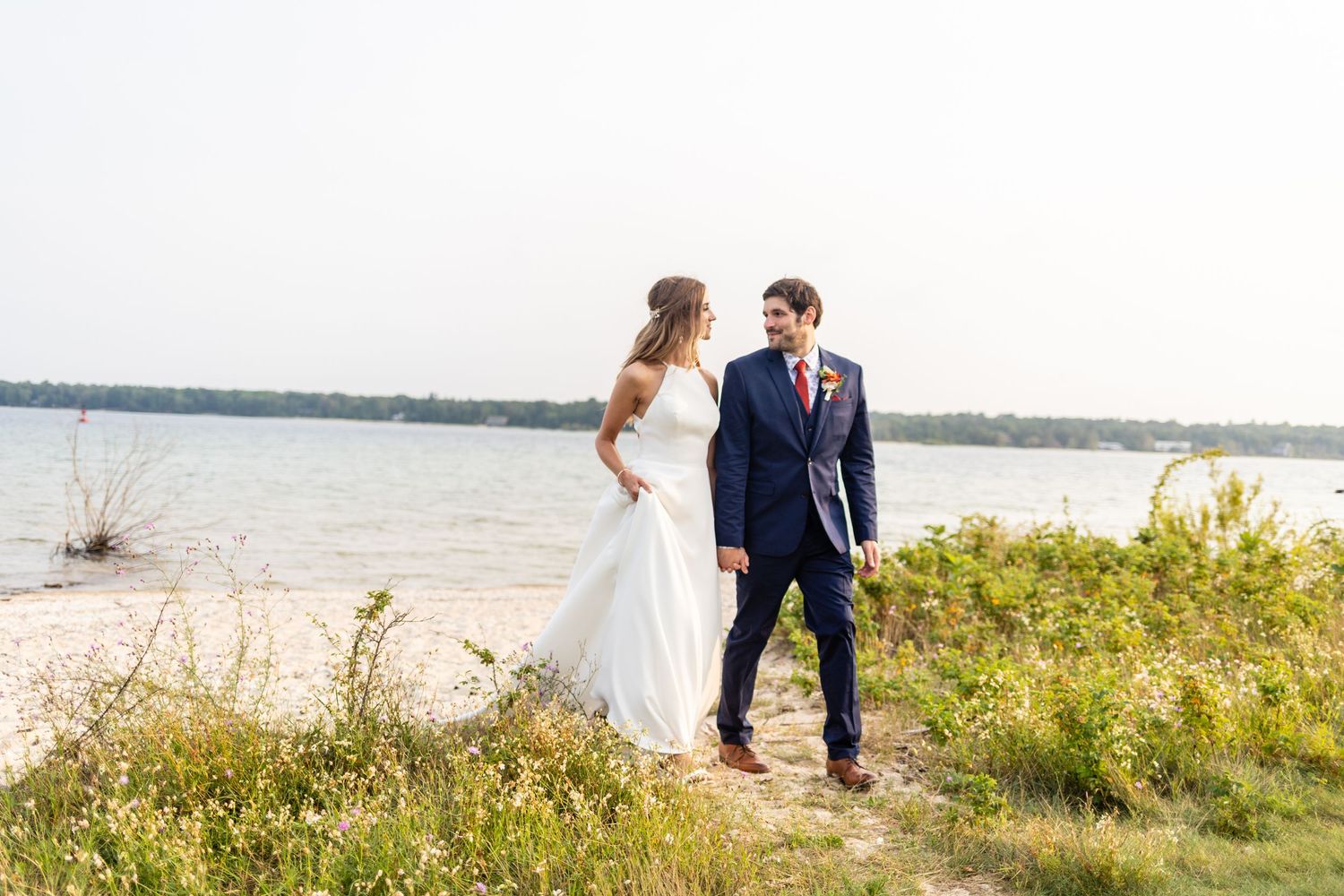 A bride and groom stand together on a sandy lakeside shore on their wedding day.