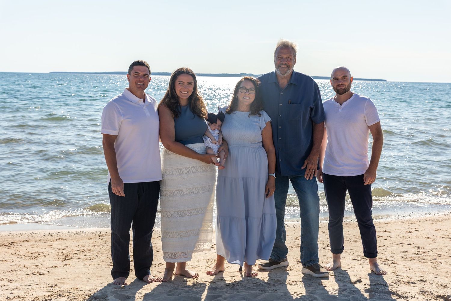 Family of five posing together on a sandy beach with calm water, one member holding a small dog.