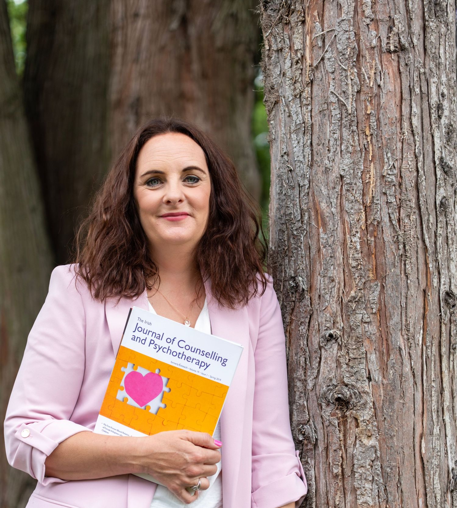 A person in a pink blazer stands by a tree trunk while holding an orange book.