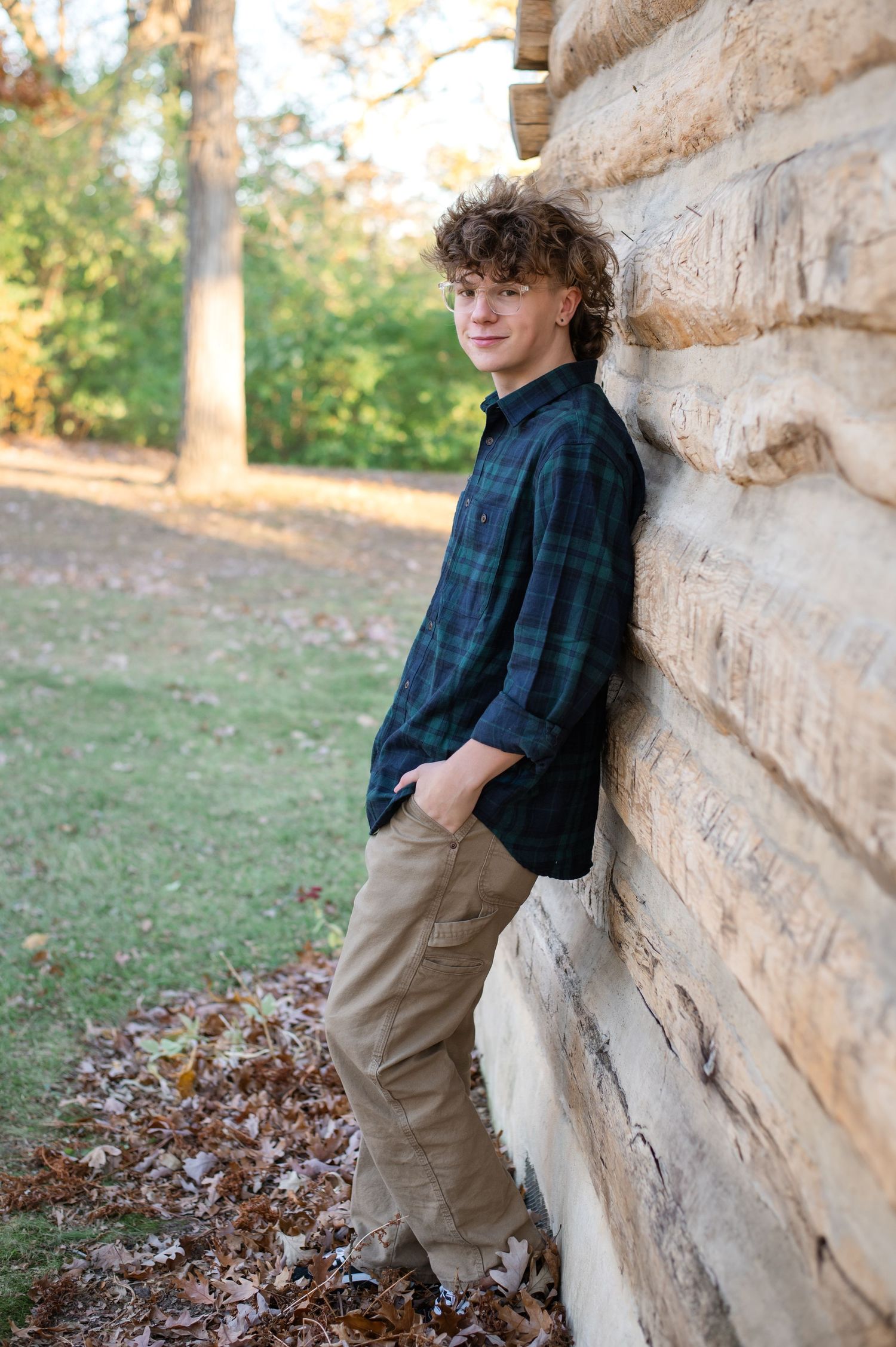 Young person in plaid shirt and khakis leans against stone wall outdoors during autumn season.