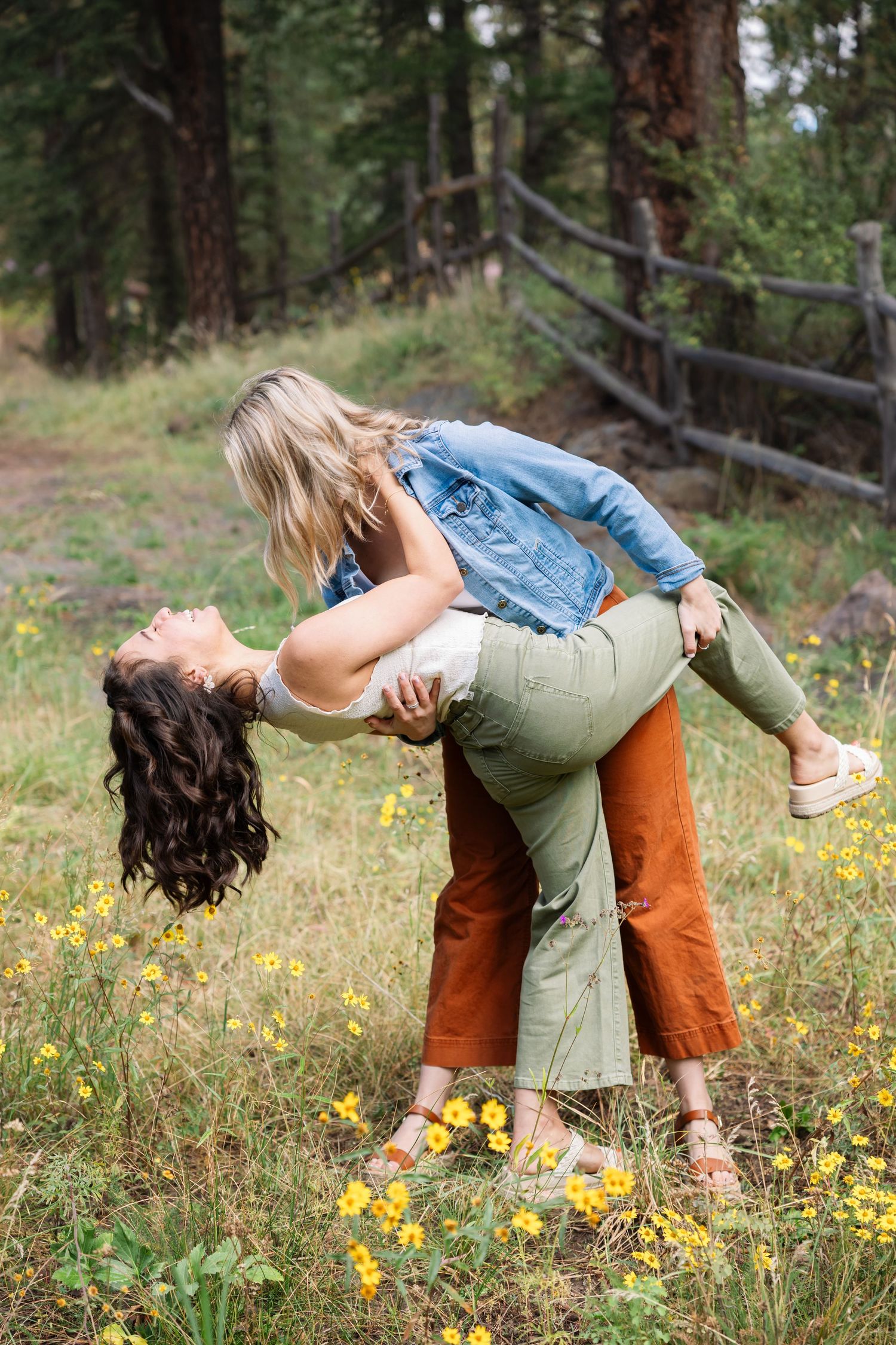 A couple dances playfully in a meadow filled with yellow wildflowers and tall pine trees in the background.