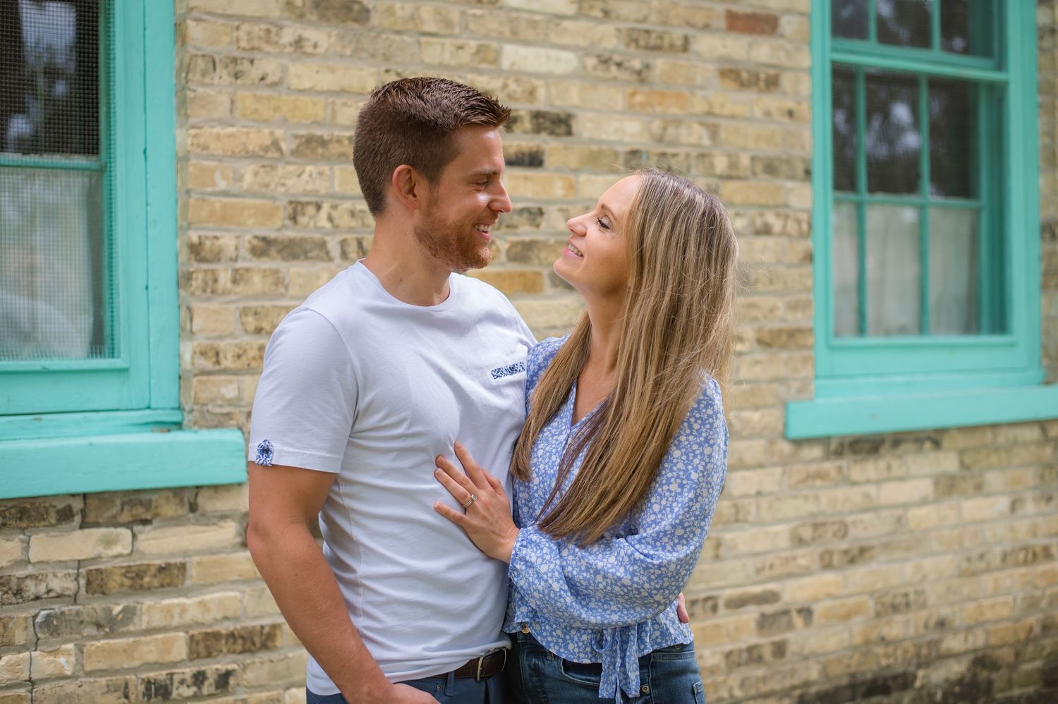 A couple shares a romantic moment together against a rustic brick wall with turquoise window frames.