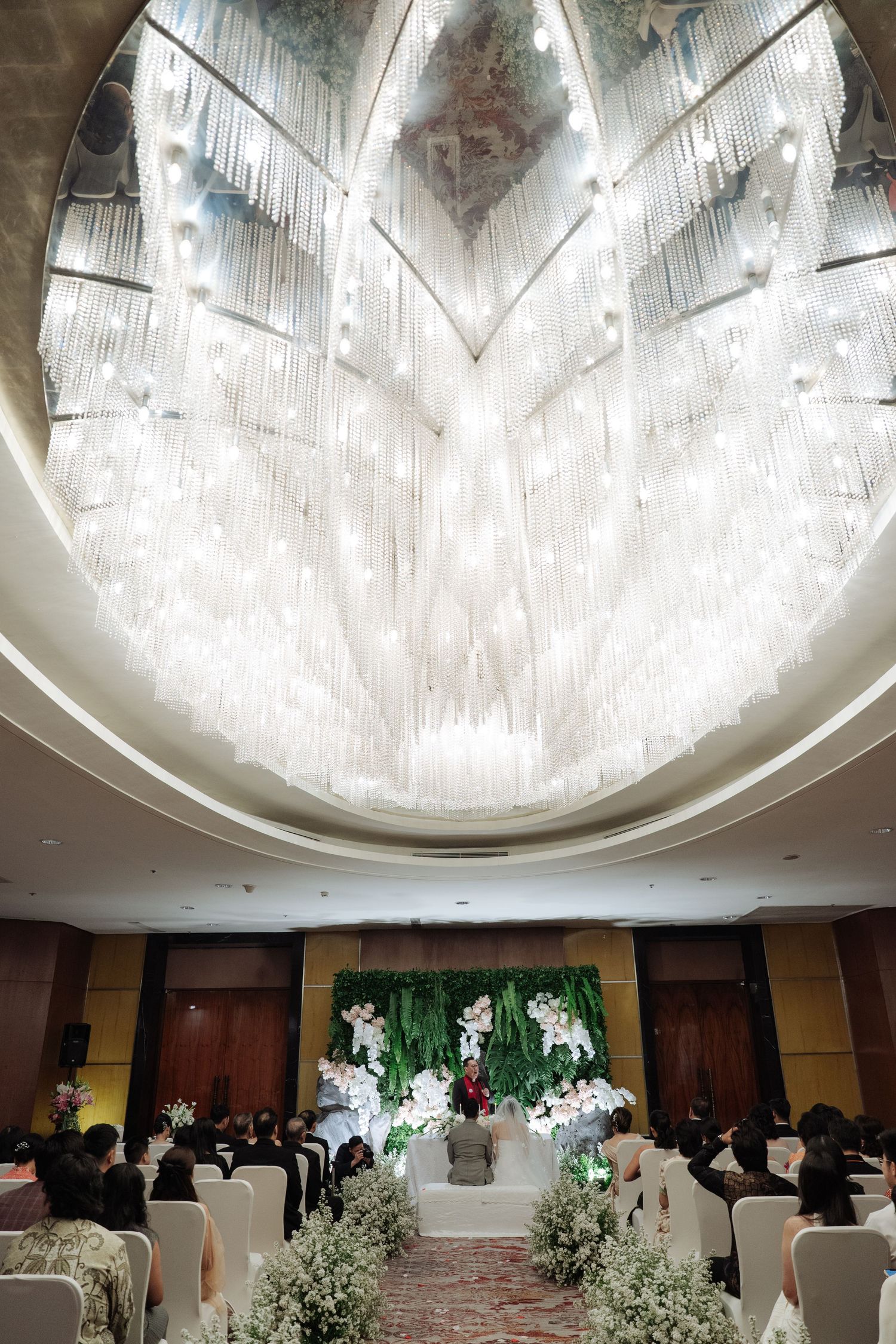 Grand wedding ceremony venue with stunning crystal chandelier ceiling and floral altar surrounded by seated guests.