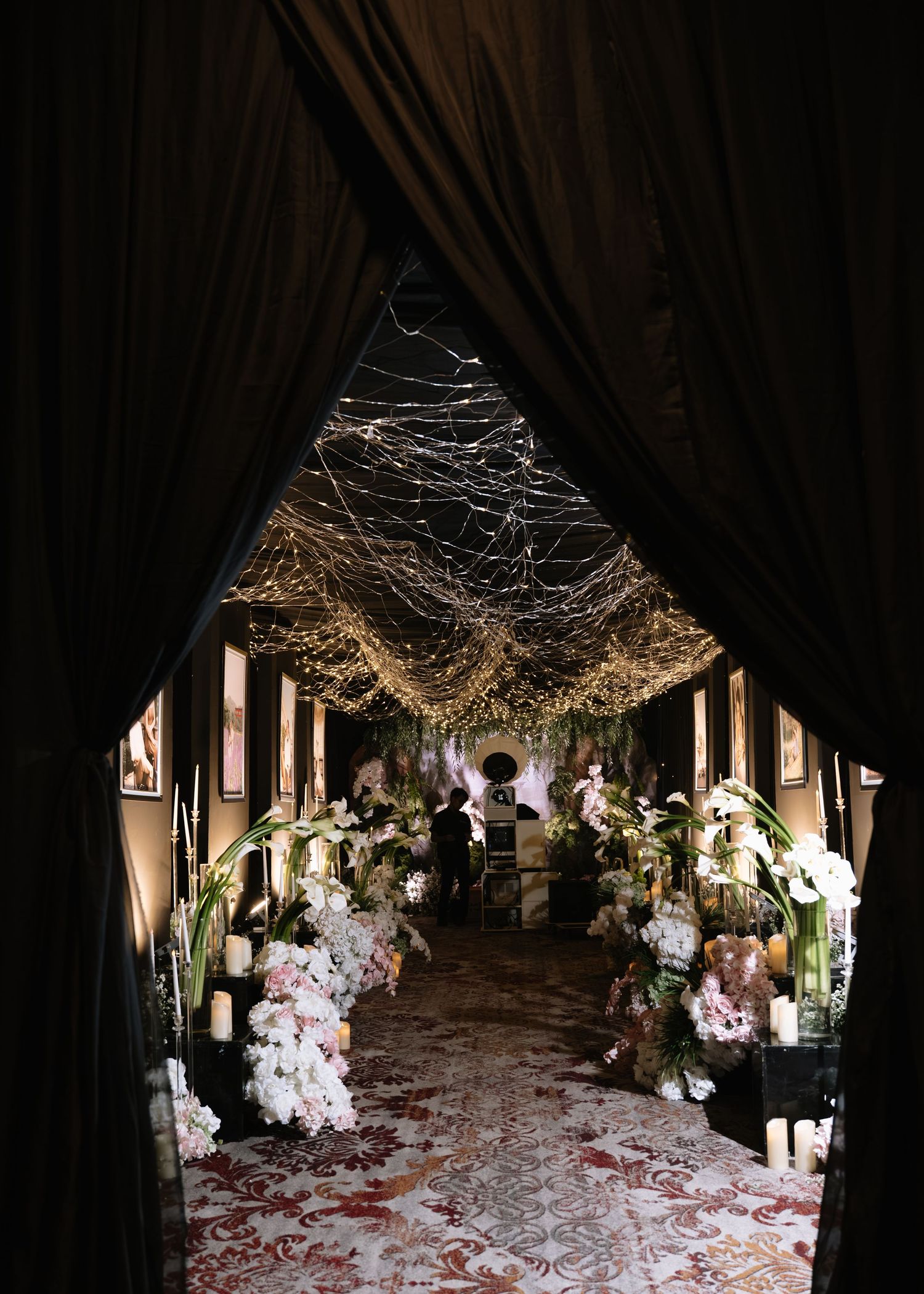 Dramatic dark wedding venue entrance with draped curtains revealing a fairy light canopy and floral aisle decor.
