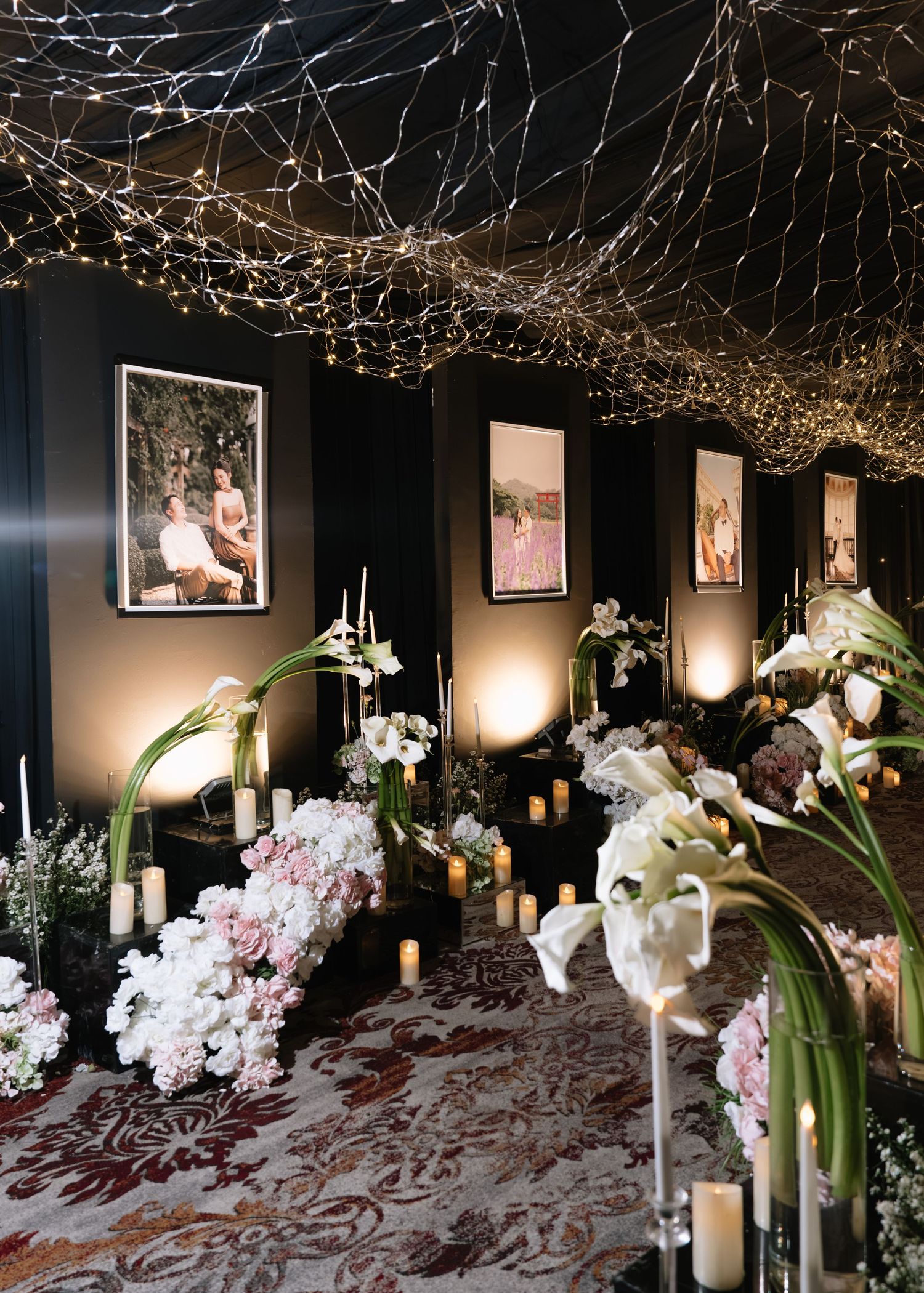Moody wedding reception space with twinkling fairy light ceiling, framed photos, candles, and white floral arrangements.