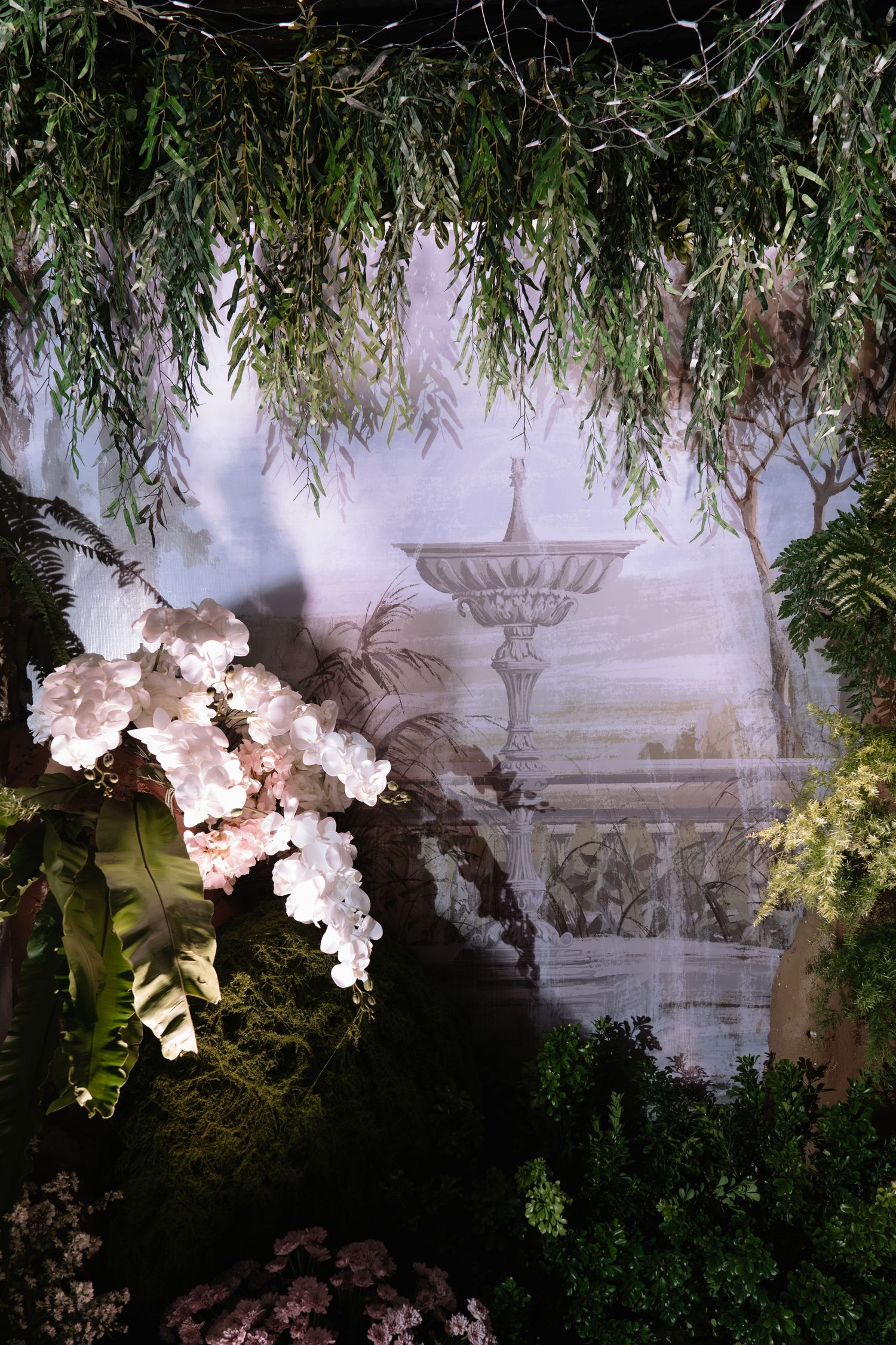 Enchanting garden-themed wedding backdrop featuring white orchids, tropical greenery, and a painted fountain mural.