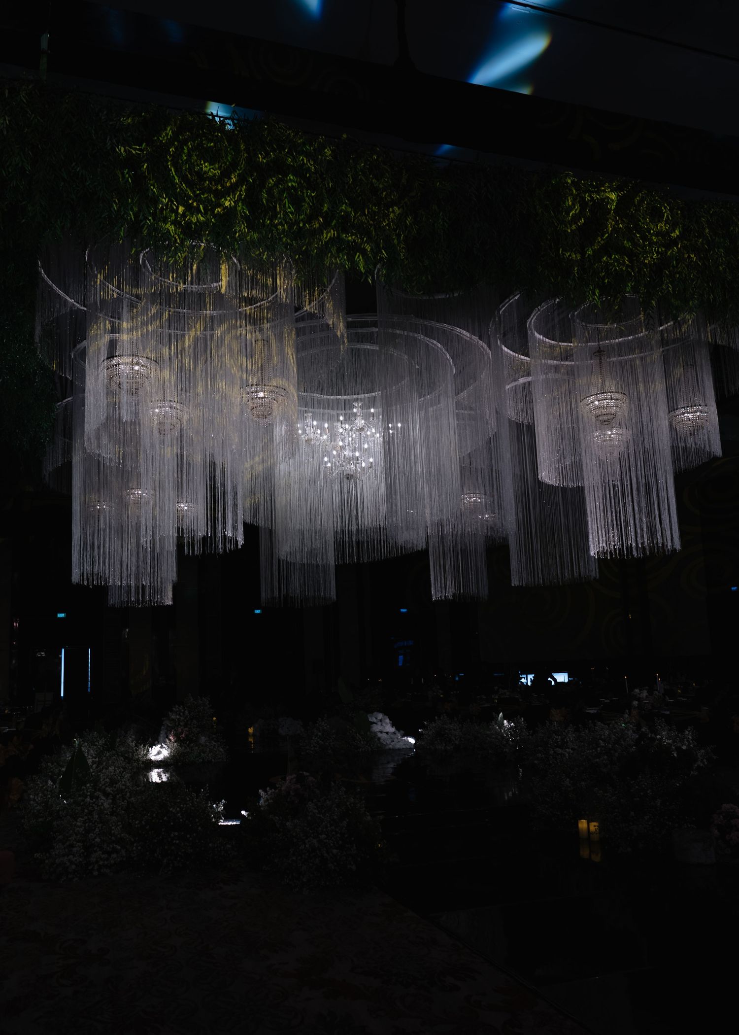 Dramatic dark wedding reception ceiling installation with cascading crystal strands and chandelier lighting above florals.