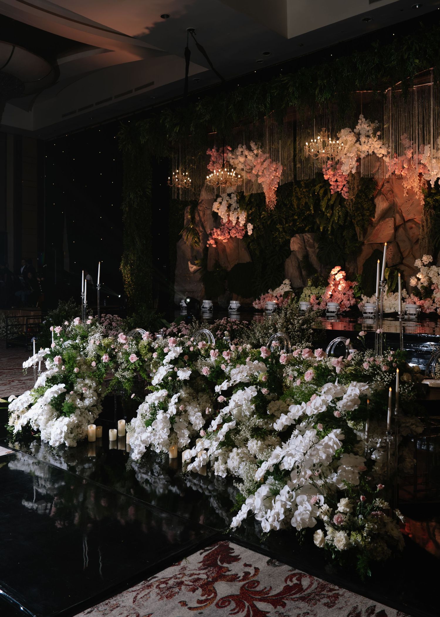 Lush white floral wedding backdrop with pink blooms, greenery, and candles in an elegant dark reception hall.