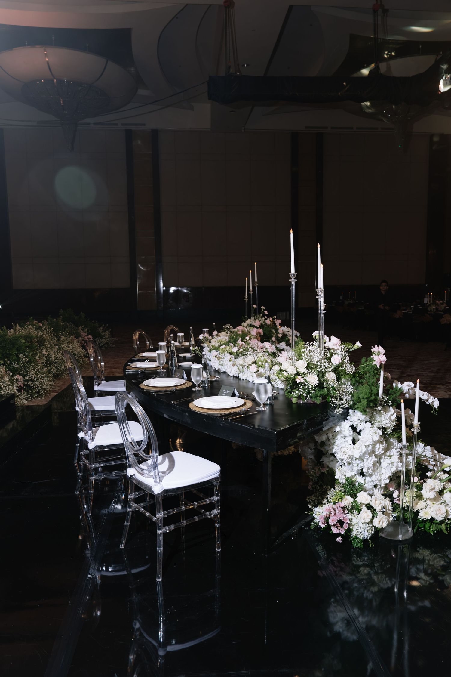 Elegant wedding reception table with ghost chairs, white florals, candles, and black table in dark ballroom.