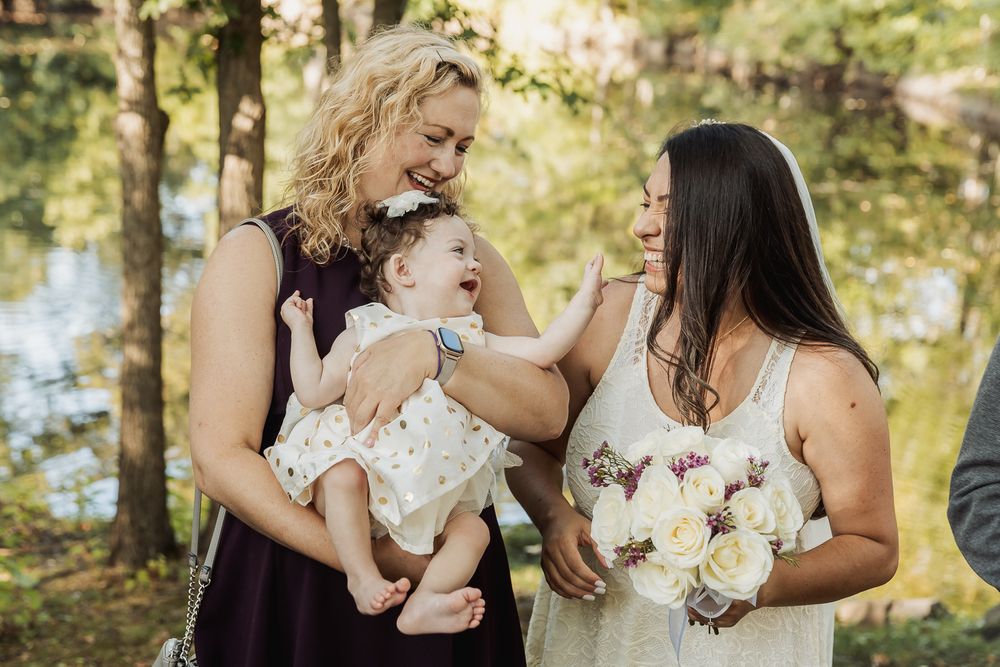 Three people share a tender moment outdoors in a natural setting with trees and water in the background.