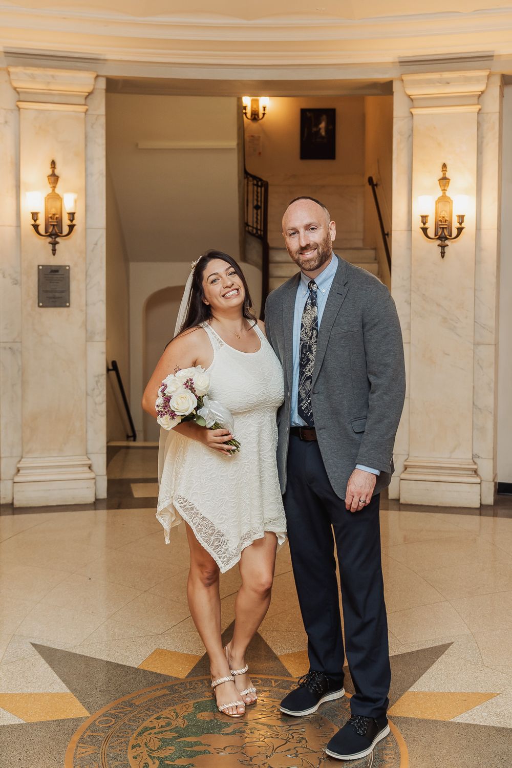 Couple poses playfully in an elegant hallway with marble columns and wall sconces.