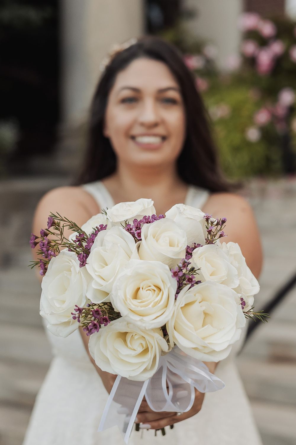 A wedding bouquet of white roses with purple accents held against a white dress.