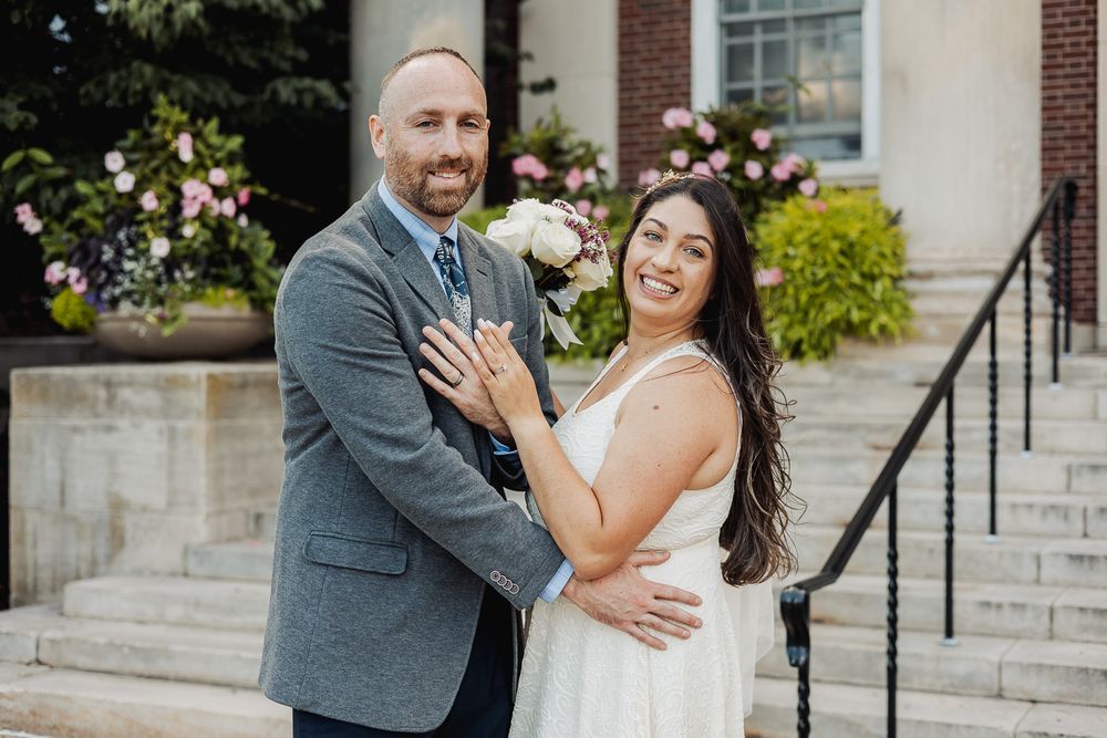 A couple in wedding attire share romantic moments in front of a brick building with blooming spring flowers.