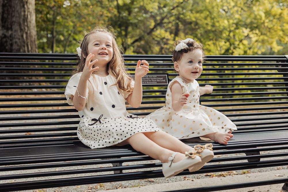 Children sit on a park bench in white dresses during a sunny outdoor photoshoot.