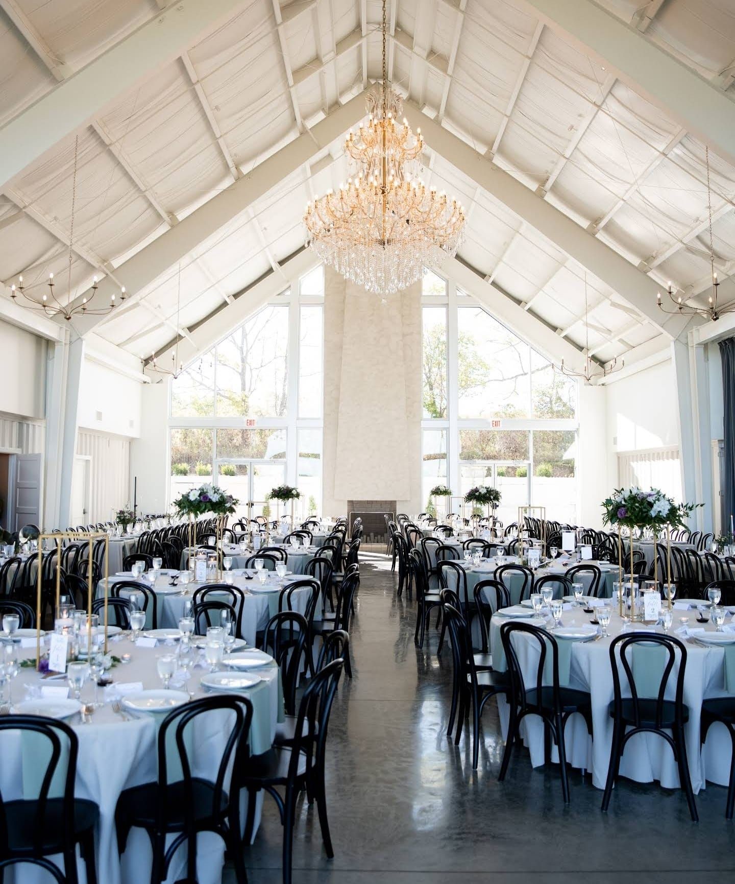 White reception hall with vaulted ceiling, chandeliers and black chairs set for wedding dinner.