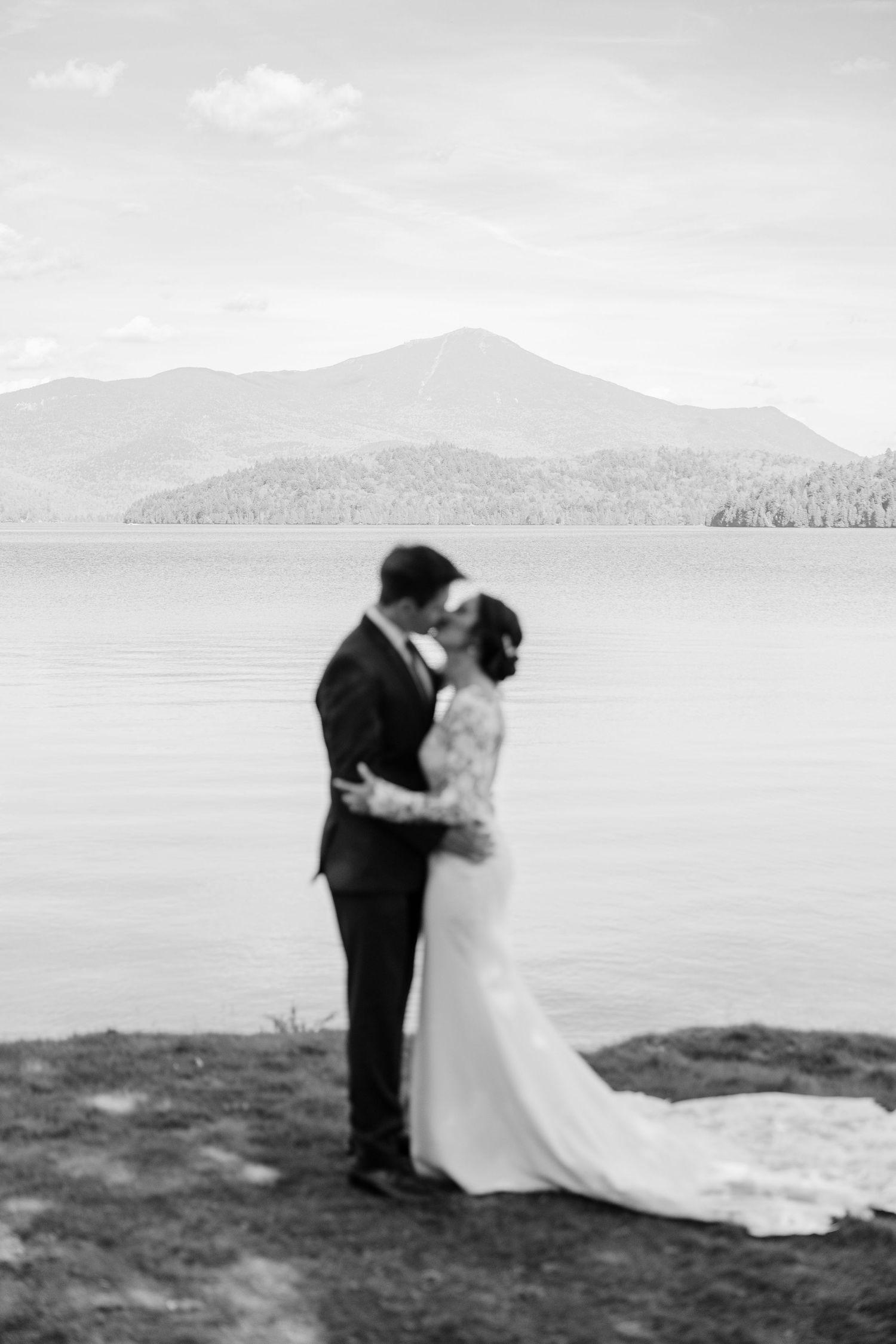 A couple in wedding attire embraces on a scenic cliffside overlooking a serene lake with mountains in the background.