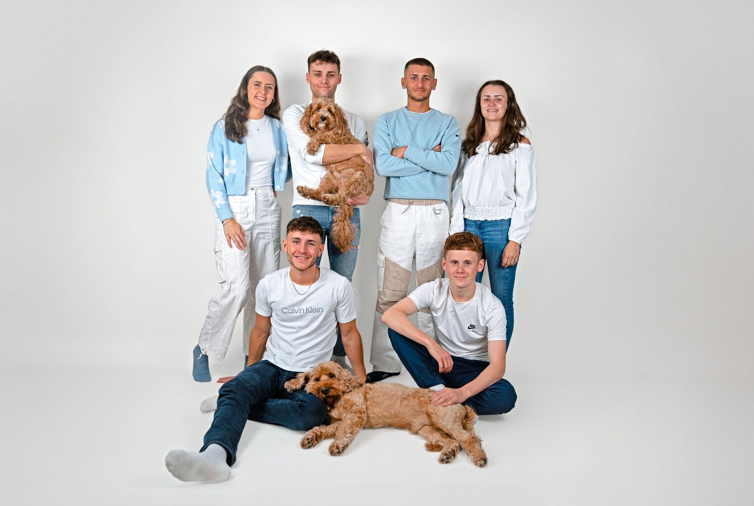 Group of six young people in white and blue outfits posing with two golden doodle dogs in a studio.
