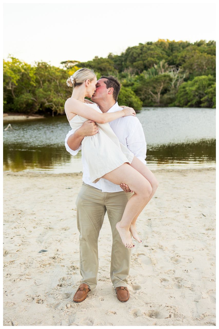 Romantic couple shares tender moments on a sandy beach beside a tranquil lake during golden hour.