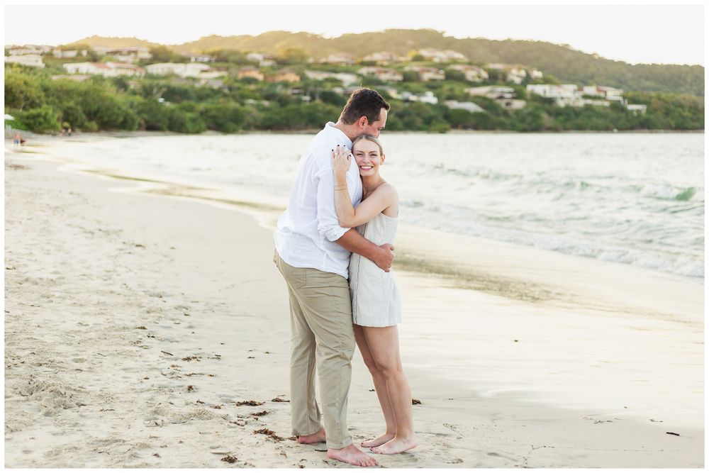 A couple shares a romantic moment on a scenic beach with rolling hills in the background during sunset.