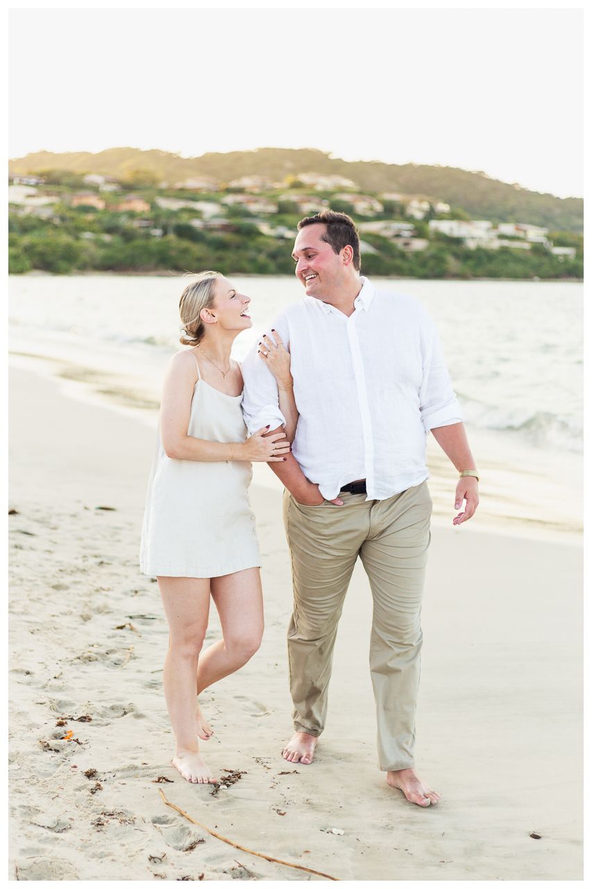 A couple wearing white and khaki outfits walking along a scenic beach at sunset with mountains in the background.