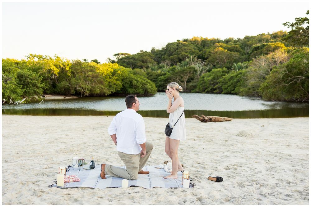 A romantic beach picnic setup at sunset with blankets and refreshments laid out on golden sand near the water.