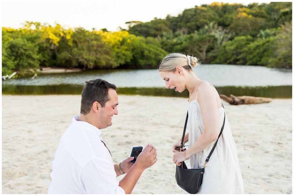 A romantic proposal moment unfolds on a scenic beach with water and trees in the background.