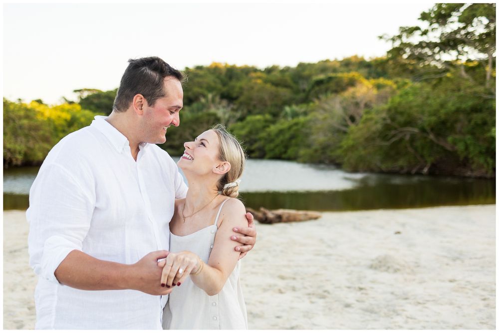 Romantic couple shares tender moments on a sandy beach with lush greenery in the background at sunset.