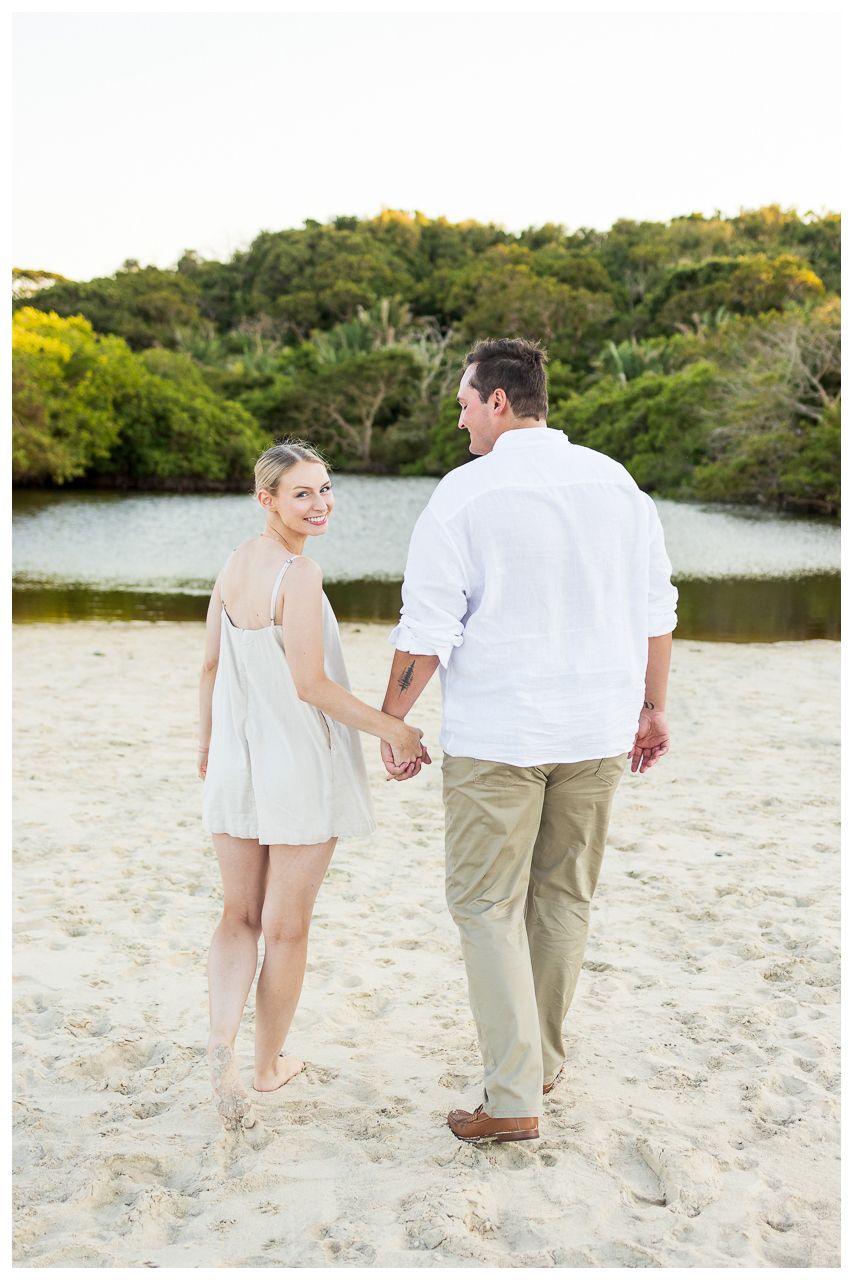 Couple walking hand in hand along a scenic beach path surrounded by coastal vegetation.