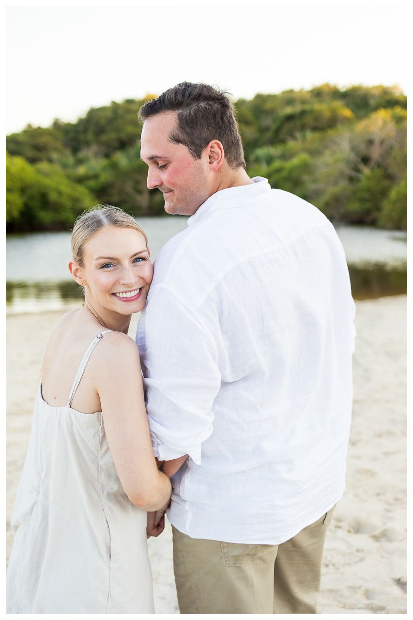 Couple in casual beach attire embracing and smiling on sandy shore with water and greenery in background.