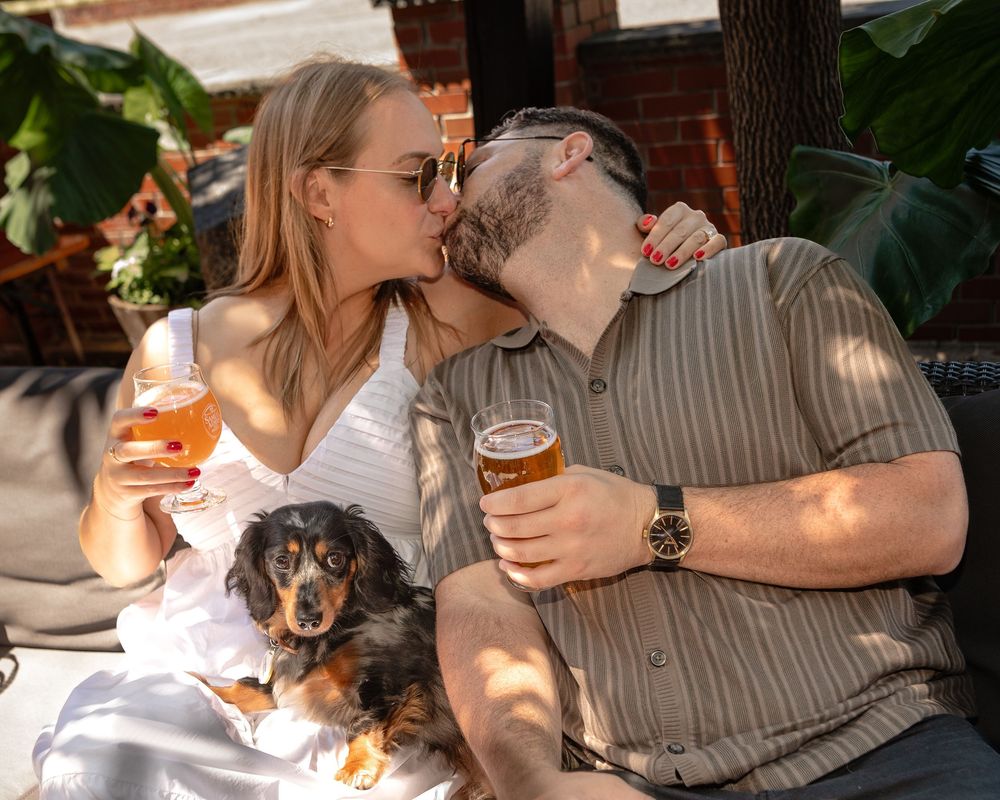 Couple enjoying drinks at outdoor patio with small dachshund dog sitting between them in dappled sunlight.
