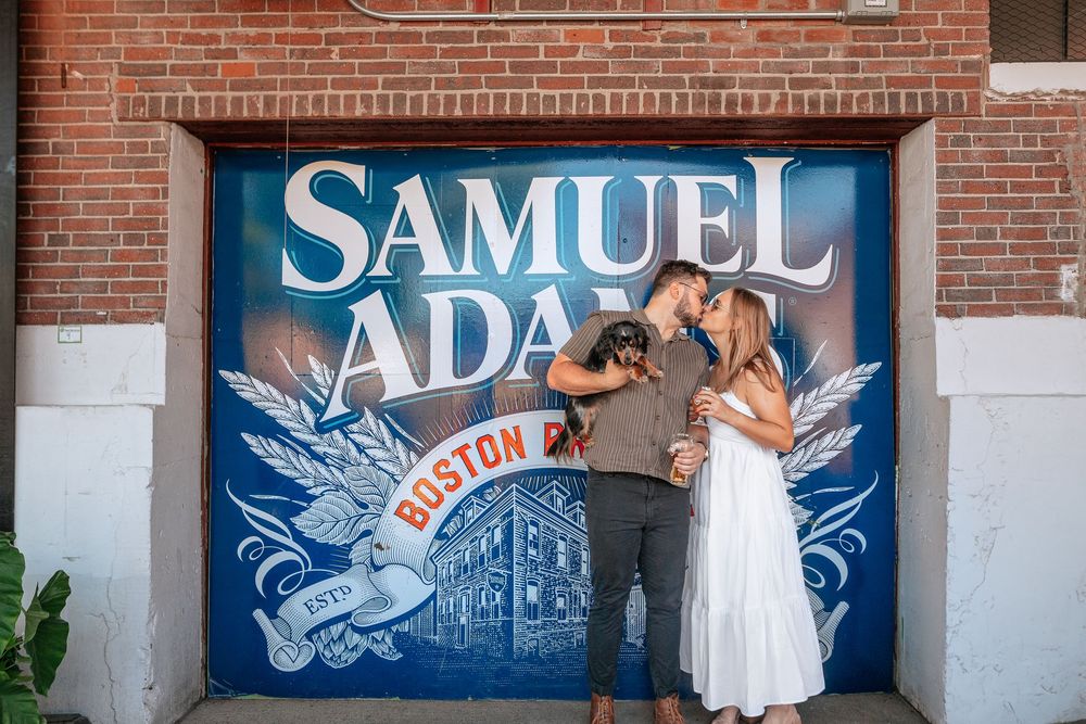 Two people pose in front of a large blue Samuel Adams brewery sign with decorative wings on a brick wall.