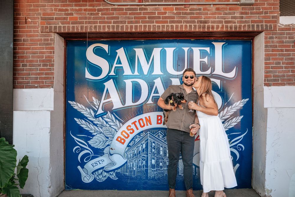 A couple poses in front of a blue Samuel Adams Boston Brewery mural painted on brick wall.