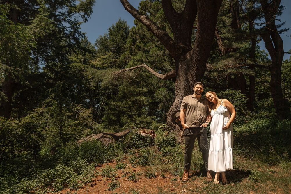 A couple poses together in a wooded outdoor setting with pine trees and natural sunlight filtering through the branches.