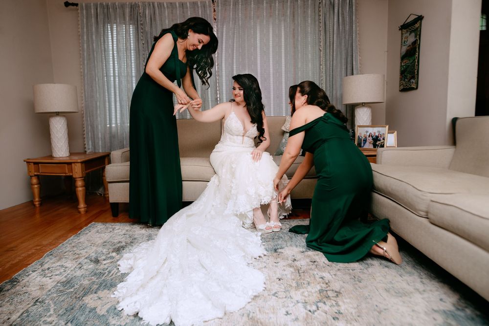Bridesmaids in emerald green dresses help adjust a bride's flowing lace wedding gown in a sunlit living room.