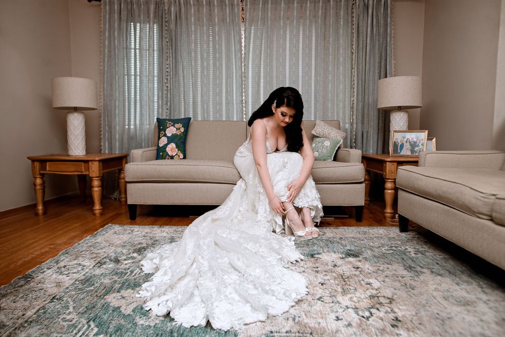 A bride in a flowing white lace gown adjusts her shoe while sitting on an elegant gray patterned rug in a living room.