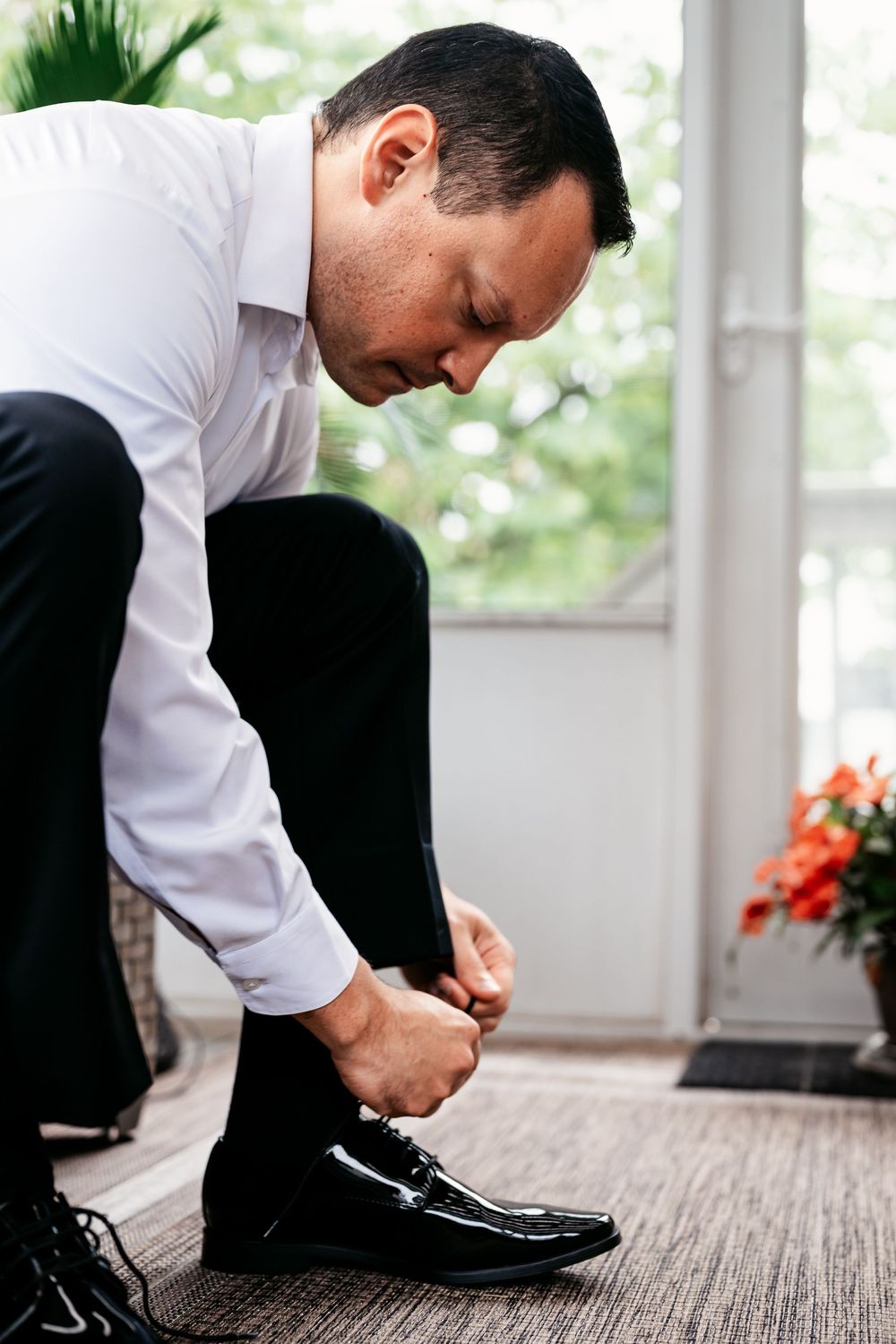 Person in formal attire adjusts their dress shoes while getting ready on a carpeted floor near a window.