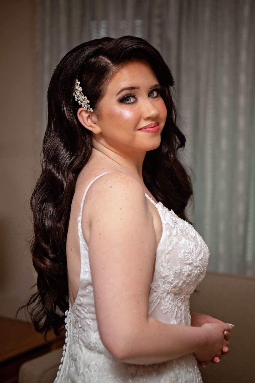 A bride in a white lace wedding dress with dark curled hair poses for portraits against a light background.