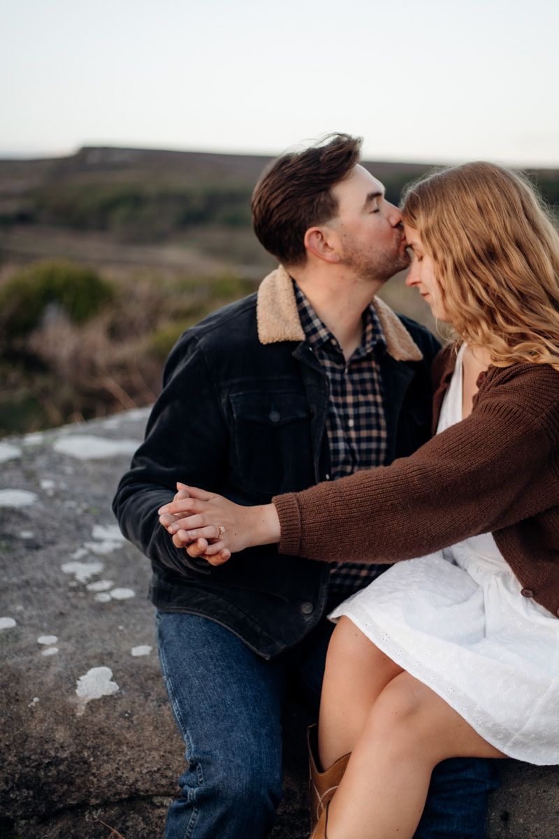 A couple shares a tender moment, the man kissing the woman's forehead while holding hands outdoors.