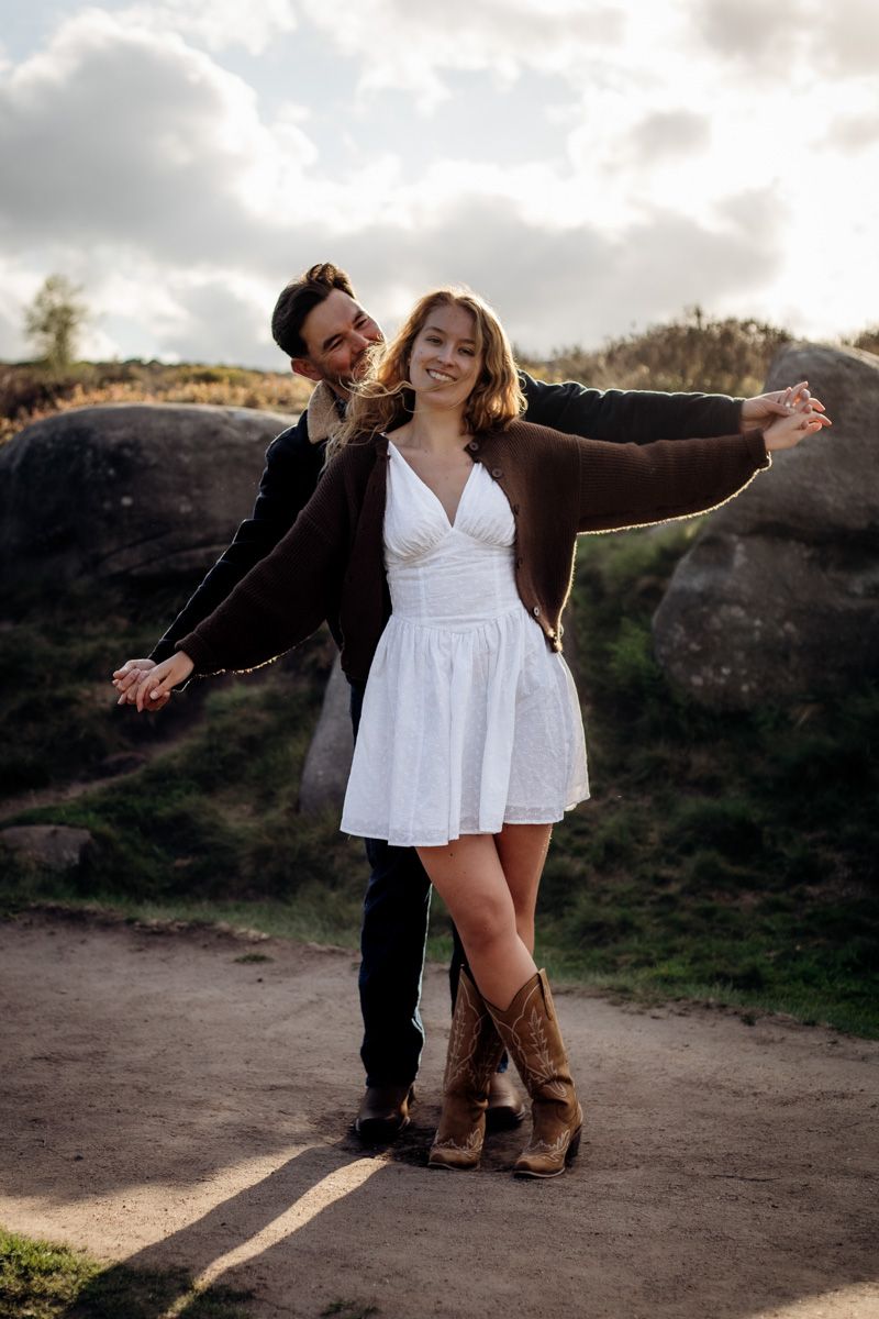 Couple dancing outdoors, woman in white dress and cowboy boots with arms outstretched, man in black holding her hands.