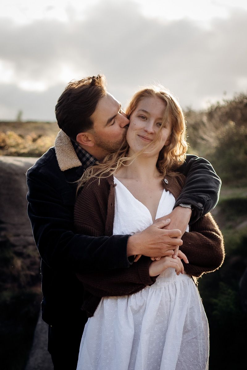 Couple embracing outdoors, man kissing woman's cheek, she wears white dress and brown cardigan, cloudy sky background.