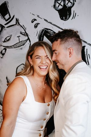 A laughing couple in white outfits poses together in front of a black and white graffiti mural wall.